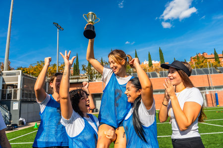 Happy young women soccer players celebrating victory on a sunny outdoor field, lifting the championship trophy together with smiles, teamwork and energetic joy after the winの写真素材