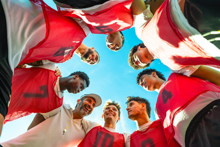 Coach and young diverse male soccer players forming a huddle, discussing strategy and fostering collaboration, looking down at them from below against a bright blue skyの写真素材