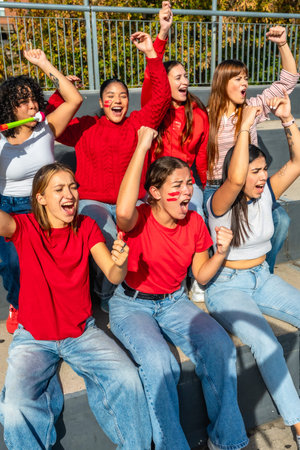 Young women sitting on stadium bleachers, cheering loudly with raised arms and painted faces, expressing excitement and support for their favorite team during a matchの写真素材