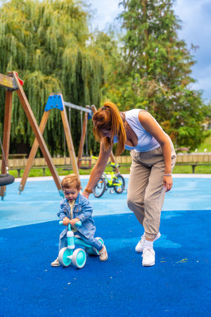 Young mother helping her toddler son learn to ride a balance bike on a playground's soft blue surface, promoting early childhood development and family bondingの写真素材