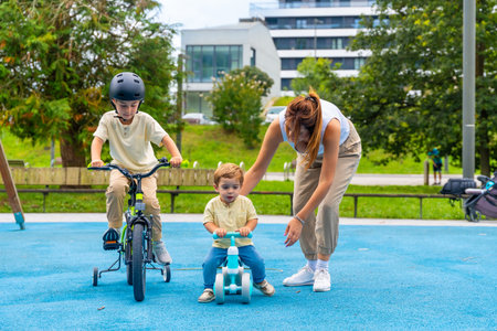 Young mother assisting two children riding bikes in a sunny playground, with the older boy on a bicycle with training wheels and the younger on a balance bikeの写真素材