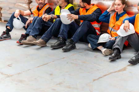 Group of diverse warehouse workers sitting on the floor taking a break, feeling exhausted after a long shift, wearing safety workwear in a logistics warehouseの写真素材