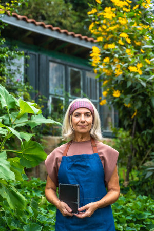 Senior woman wearing an apron and a headband, standing in a lush green garden while holding a modern tablet, representing the blend of tradition and technology in gardeningの写真素材
