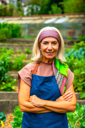 Senior woman wearing an apron and headscarf, standing with arms crossed in a lush community vegetable garden, smiling and looking directly at the viewerの写真素材
