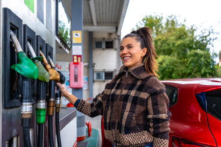 Woman smiling and wearing a protective glove while selecting a fuel pump to refuel her red car at a gas station, showcasing daily tasks and vehicle maintenanceの写真素材