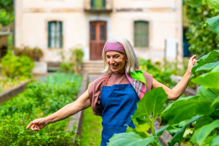Senior woman with gray hair and colorful headband smiling while standing in her flourishing home vegetable garden, appreciating sustainable and healthy livingの写真素材