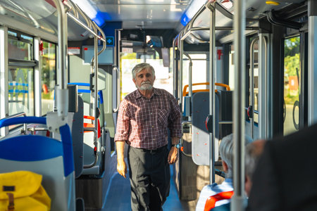 Senior man with a beard and plaid shirt standing in the aisle of a public bus, commuting during the day, with other passengers seated and windows bringing in natural lightの写真素材