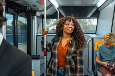 Woman with afro hair and eyeglasses standing and holding a handrail on a public transport bus, smiling while traveling, engaging in daily commute or urban journeyの写真素材