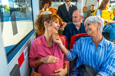Pregnant woman and senior woman are smiling and talking while traveling on a public bus, sharing a happy moment together in a diverse group of passengersの写真素材