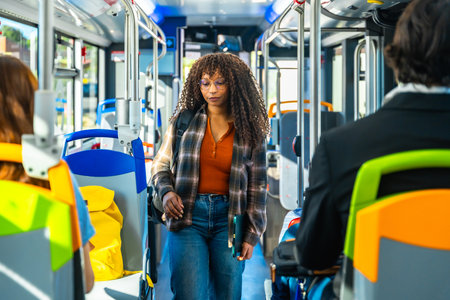Young woman passenger with long curly hair and glasses walking along the aisle of a public bus, carrying a laptop and a backpack, surrounded by vibrant seat colorsの写真素材