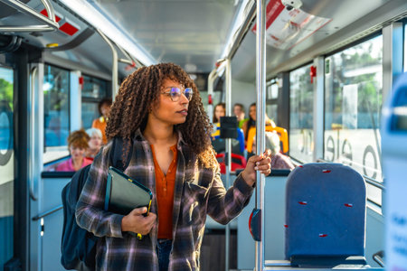 Young woman with curly hair and glasses stands inside a city bus holding a pole, wearing a backpack while commuting through urban streets during a daytime journeyの写真素材