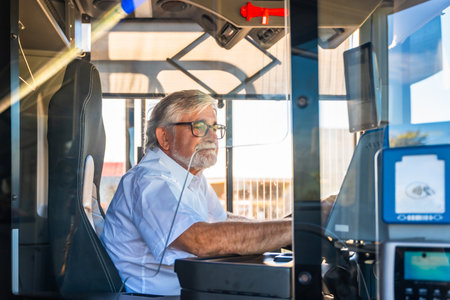 Senior bus driver with eyeglasses and white shirt focused on steering from the cabin, demonstrating dedication and experience in urban public transportation during daytime commuteの写真素材