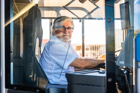 Senior man with a white beard and glasses wearing a shirt, sitting at the driver's seat of a modern bus during daylight, smiling and working in public transportationの写真素材