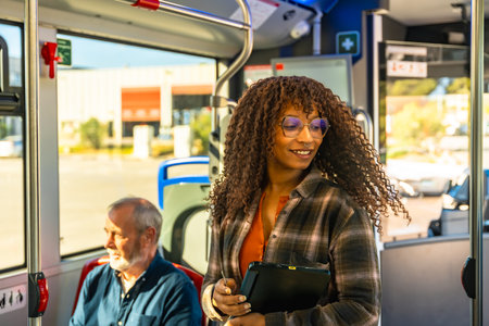 Woman with glasses smiles while standing on a city bus, holding a digital tablet among commuters during a bright daily commute, modern, eco friendly urban travelの写真素材