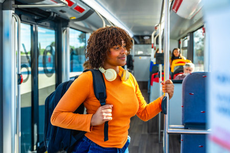 Young black woman with headphones and backpack standing in a public transport bus, holding a handrail and smiling, representing urban lifestyle and daily commuteの写真素材