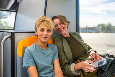 Mother and son sitting inside a bus, sharing a moment while traveling using public transport, the mother scrolling on a smartphone and the son looking at the viewerの写真素材