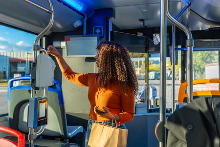 Young woman with curly hair validating her public transport ticket on a bus, holding a smartphone and a paper shopping bag, commuting morning or eveningの写真素材