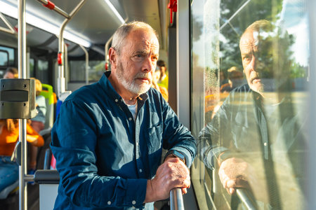 Senior bearded man standing on a city bus, holding a handrail and gazing out the window, bathed in...