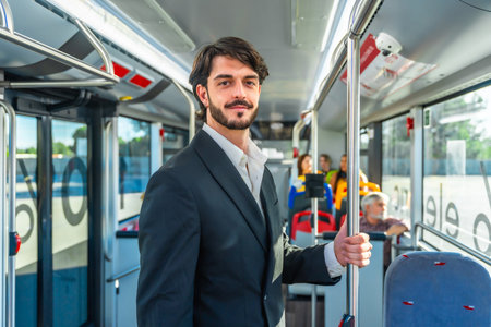Young businessman traveling by bus, holding a pole and smiling while looking at the camera, with other passengers in the background, representing daily commute and urban lifestyleの写真素材