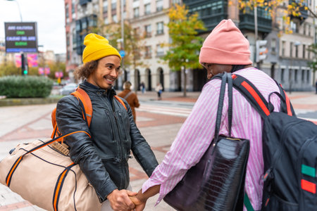 Multiethnic couple holding hands, beanies and backpacks, smiling as they walk through a vibrant city street with luggage, enjoying an autumn urban travel momentの写真素材