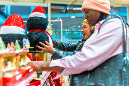 Diverse couple browsing holiday decorations in a store, a man looking surprised and excited while holding a penguin decoration, creating a joyful moment during Christmas shoppingの写真素材