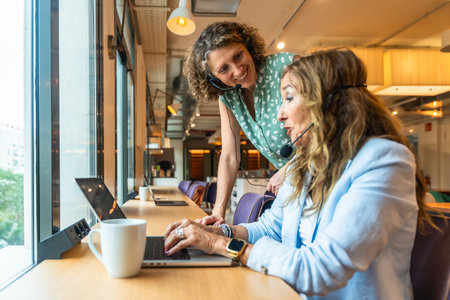 Two women wearing headsets are working together at a desk by a window, with one colleague providing assistance and guidance to the other using a laptop in a coworking spaceの写真素材
