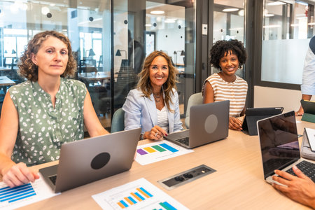 Diverse businesswomen are smiling at the camera, working with laptops and analyzing data graphs while collaborating around a table in a modern office meeting roomの写真素材
