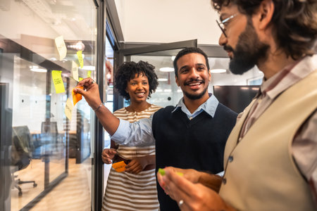 Diverse business colleagues collaborating on a project, smiling and applying colorful sticky notes to a glass wall during a creative brainstorming session in a modern officeの写真素材