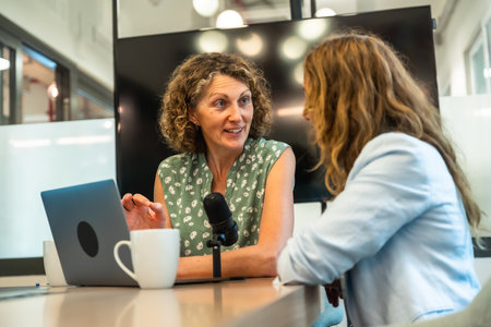 Two professional women collaborating on a podcast, recording a conversation with a microphone and laptop in a contemporary coworking space, focusing on content creation and communicationの写真素材