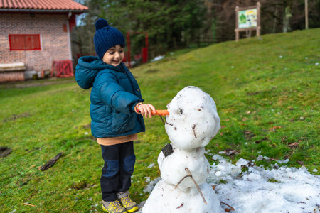 Young boy smiling and decorating a melting snowman with a carrot nose, wearing a warm hat and jacket while playing in a snowy patch on a green grassy hillの写真素材