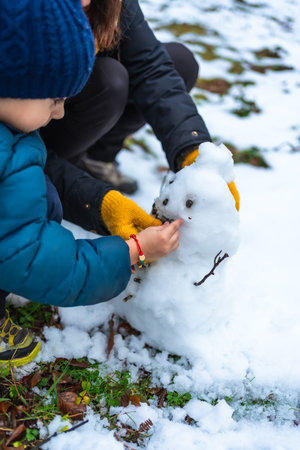 Young boy and mother collaborating to build a small snowman, carefully adding details like eyes and mouth with natural elements in a snowy outdoor setting during winterの写真素材