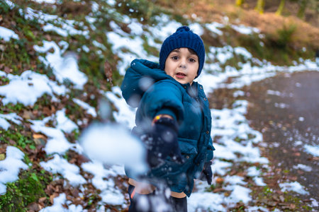Young boy wearing a blue knitted hat and warm winter jacket, actively playing and throwing a snowball directly at the viewer, enjoying the fresh snow in a natural forest settingの写真素材