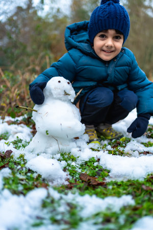 Smiling young boy crouching outdoors, happily creating a small snowman on a patch of grass lightly covered with fresh winter snow, enjoying childhood fun and creativityの写真素材