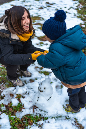 Happy mother and young son build a small snowman outdoors, laughing and bonding in warm parkas, beanies and scarves while enjoying playful winter fun and cozy family momentsの写真素材