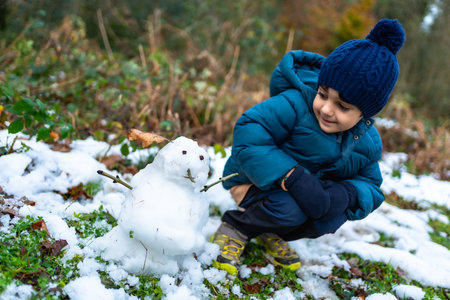 Young boy kneeling on snowy ground, wearing a warm coat, hat, and gloves, observing a freshly made small snowman with a happy expression during a cold winter dayの写真素材
