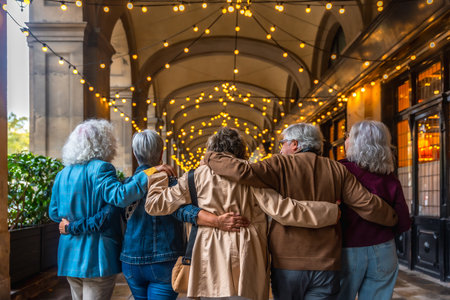 Group of diverse senior friends walking together embraced along an illuminated city archway at dusk, sharing friendship and enjoying an evening outingの写真素材