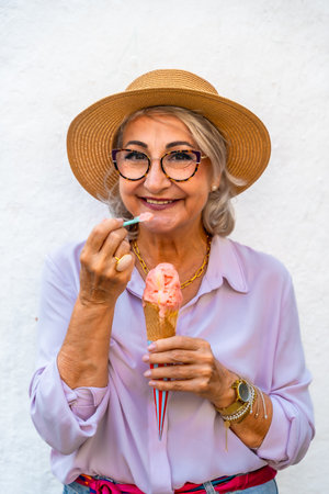 Happy senior woman wearing a straw hat and glasses smiling while eating a refreshing ice cream cone with a small spoon, celebrating moments of joy and relaxation in summerの写真素材