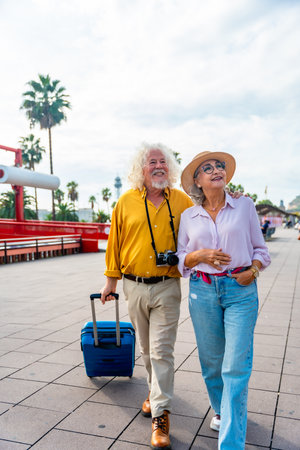 Senior couple walking on a city promenade, smiling and looking up, carrying a suitcase and a camera, enjoying their vacation and exploring new places togetherの写真素材