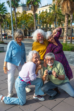 Cheerful group of senior friends traveling together, capturing fun memories with an analog camera while exploring a city street lined with palm trees on a sunny dayの写真素材