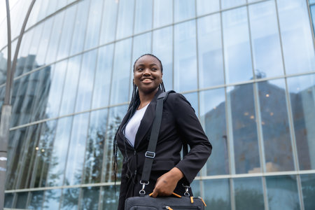 Young black businesswoman smiling and holding a laptop bag, walking with confidence outside a modern...