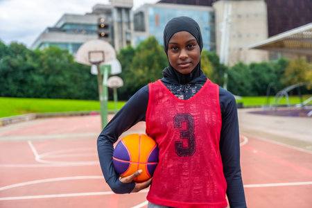 Young muslim woman wearing a hijab, long sleeve shirt, and basketball bib, holding a basketball while standing on an outdoor court, embodying confidence and athletic spiritの写真素材