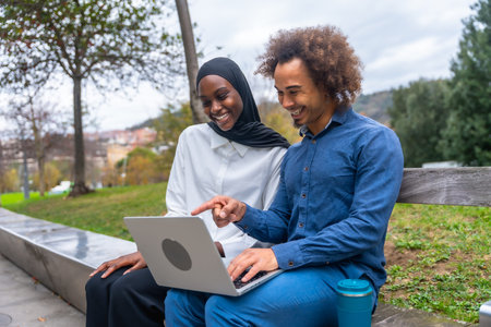 Diverse young couple sitting on a park bench, smiling and pointing at a laptop screen while sharing...