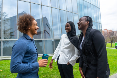 Diverse group of business colleagues, including a man holding a reusable cup and two women, laughing and smiling while standing outdoors in front of a modern glass buildingの写真素材