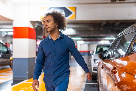 Young man with afro hair walks through a brightly lit underground parking garage, glancing right as he searches for his car among rows of vehicles and concrete columnsの写真素材