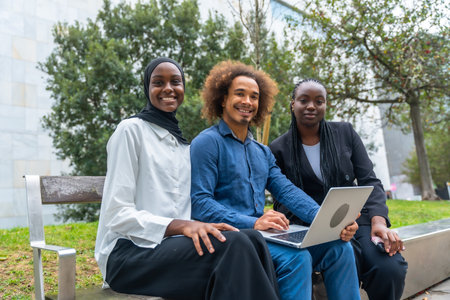 Three diverse students are smiling directly at the camera while sitting outside on a bench. Working together on a laptop. Symbolizing teamwork. Education. And modern learning in a relaxed environmentの写真素材