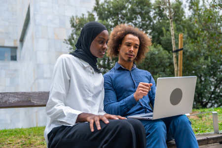 Diverse young man and woman collaborating on a laptop while sitting on a park bench, focused on work or studying together in a sunny urban green space, casual and professionalの写真素材