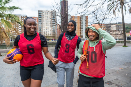 Three diverse friends, a young woman holding a basketball and two people wearing red bibs, walking...