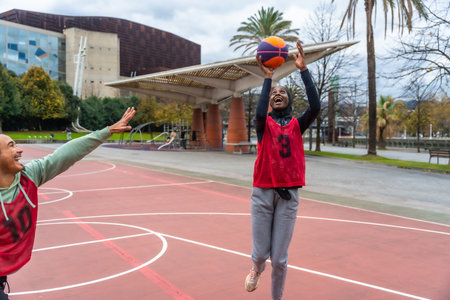 Woman in hijab smiling and shooting a basketball during a friendly game on an outdoor urban court,...