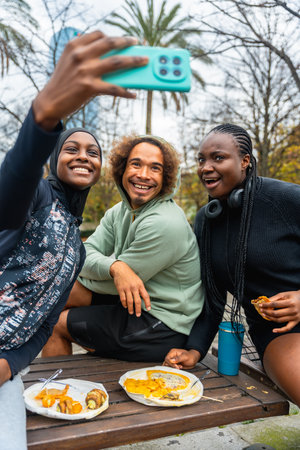 Diverse group of young adult friends smiling and laughing, holding a phone to take a selfie while enjoying a casual picnic with food and drinks on a wooden bench in an urban parkの写真素材