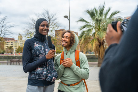 Female journalist interviewing a young, happy man with diverse background, holding a microphone while someone records a video outdoors in a city streetの写真素材